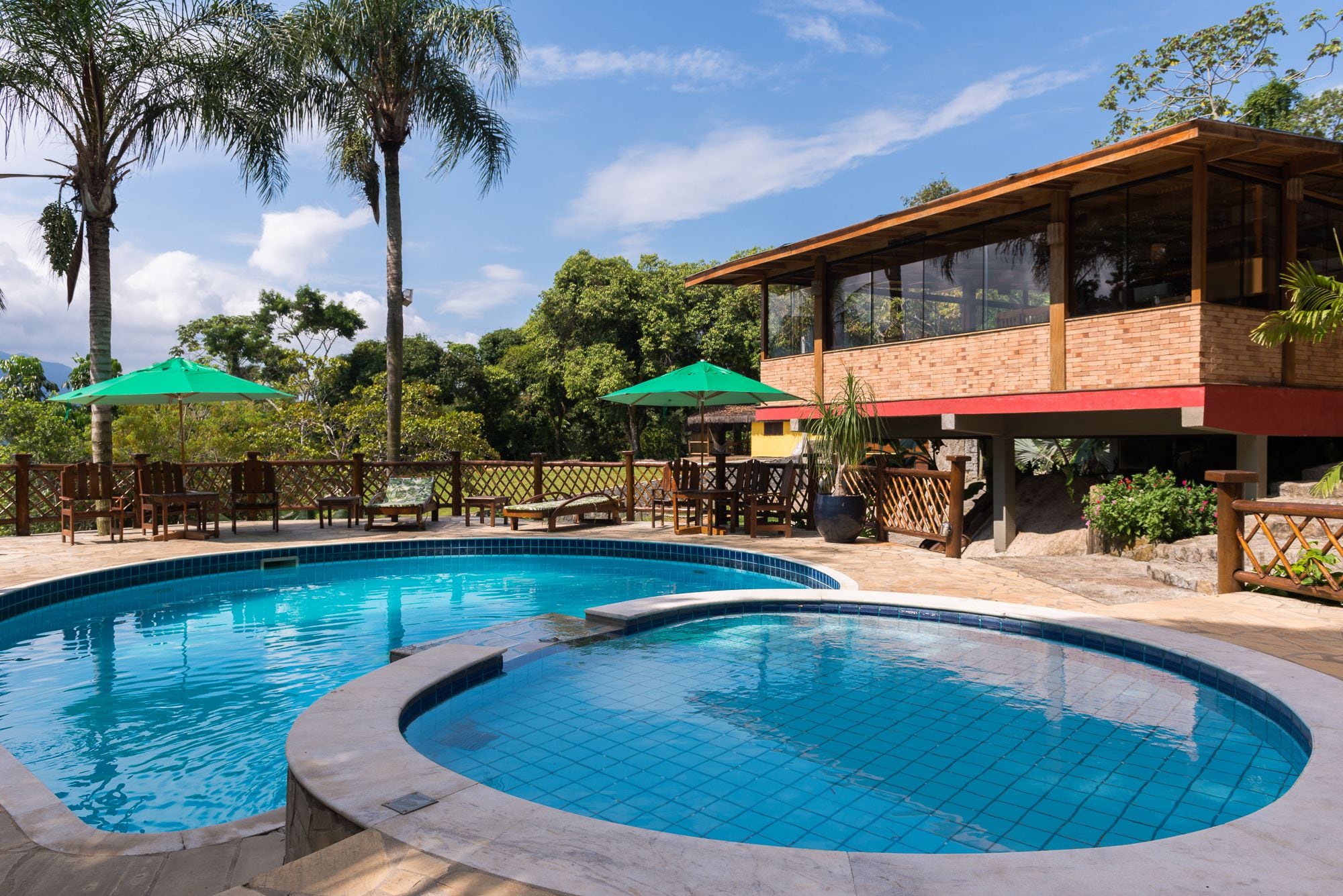 A swimming pool with a wooden deck and lounge chairs, surrounded by palm trees and a building with large windows.
