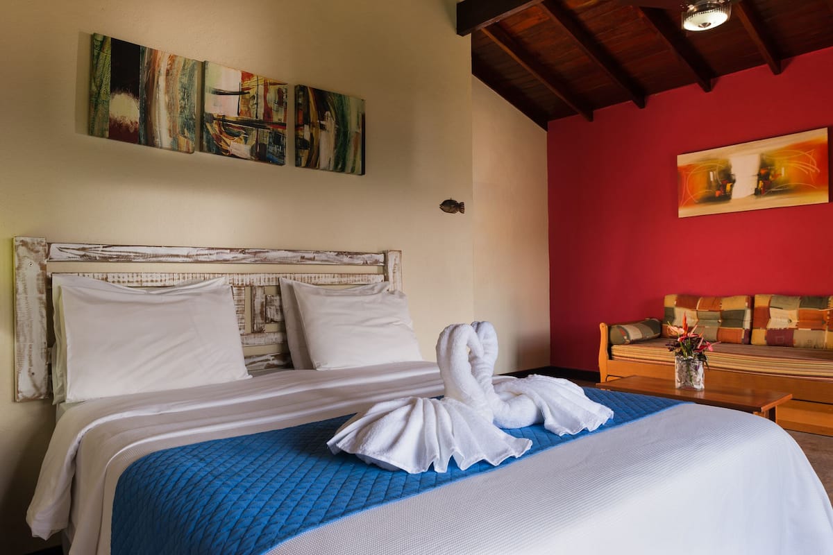 Une chambre d’hôtel avec un lit, une couvre-lit bleue matelassée, des oreillers blancs et une serviette blanche sur le lit. On y trouve une tête de lit en bois, un mur rouge et un tableau au-dessus du lit.