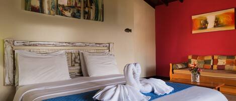 A hotel room with a bed, a blue quilted bedspread, white pillows, and a white towel on the bed. There is a wooden headboard, a red wall, and a painting above the bed.