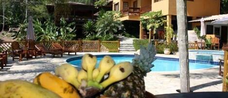 Zona de piscina con frutero que contiene plátanos y una piña, rodeada por una valla de madera y plantas tropicales.
