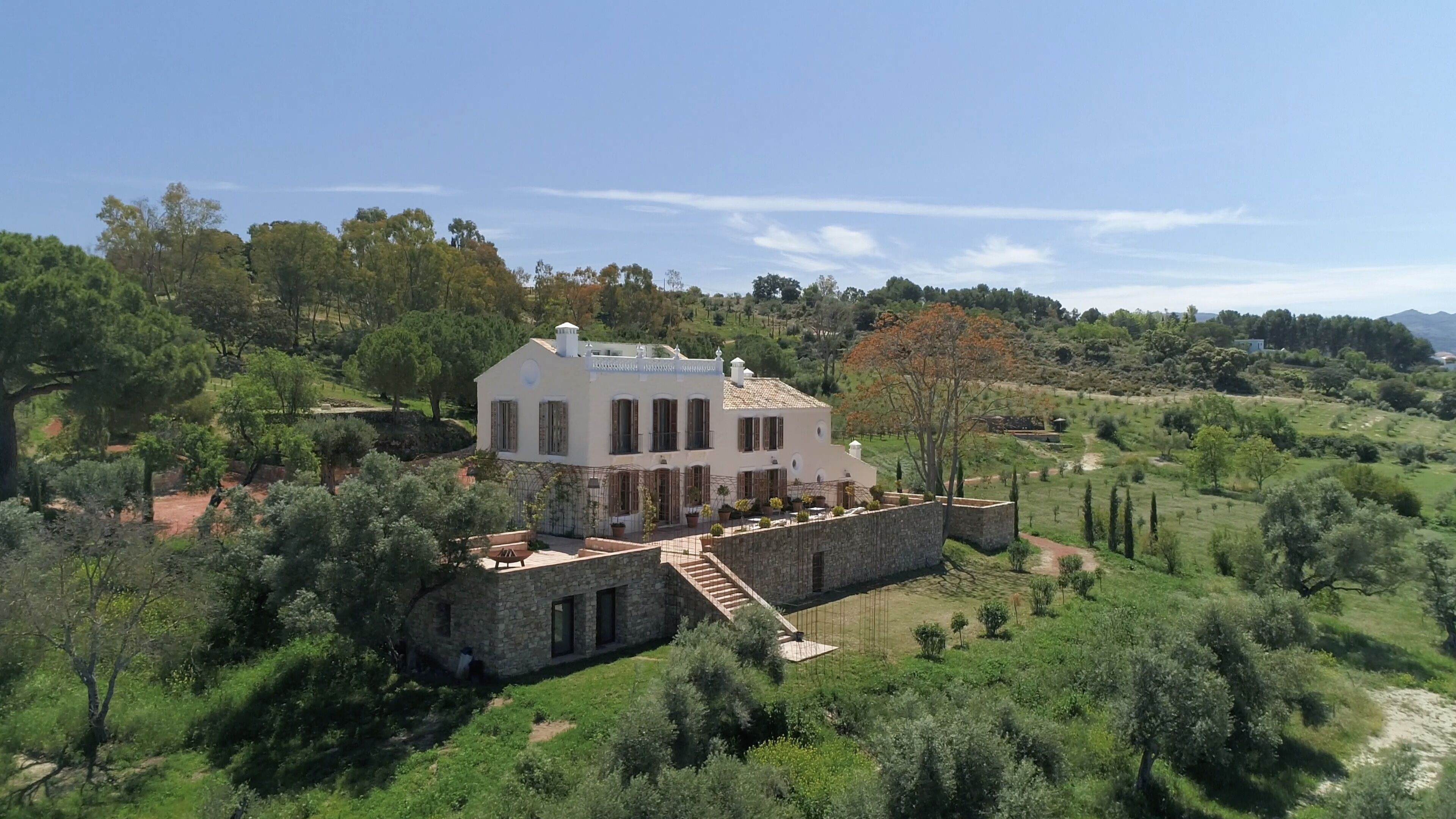 A large, two-story stone house with a stone staircase leading to the entrance, surrounded by lush greenery and trees.