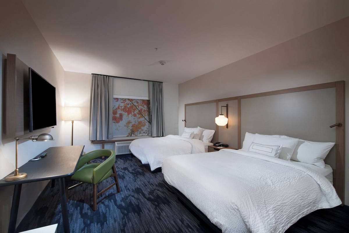 A hotel room with two beds, a desk, a chair, a TV, and a painting on the wall.