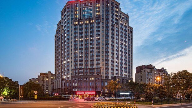 Un hôtel de grande hauteur, avec de nombreux étages, une entrée imposante agrémentée d’une enseigne lumineuse, et un environnement bien entretenu, avec des arbres et un ciel dégagé.