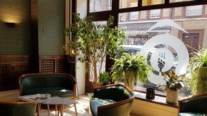 A cozy seating area with green upholstered chairs and a small table, surrounded by potted plants and a large window with a view of the street.