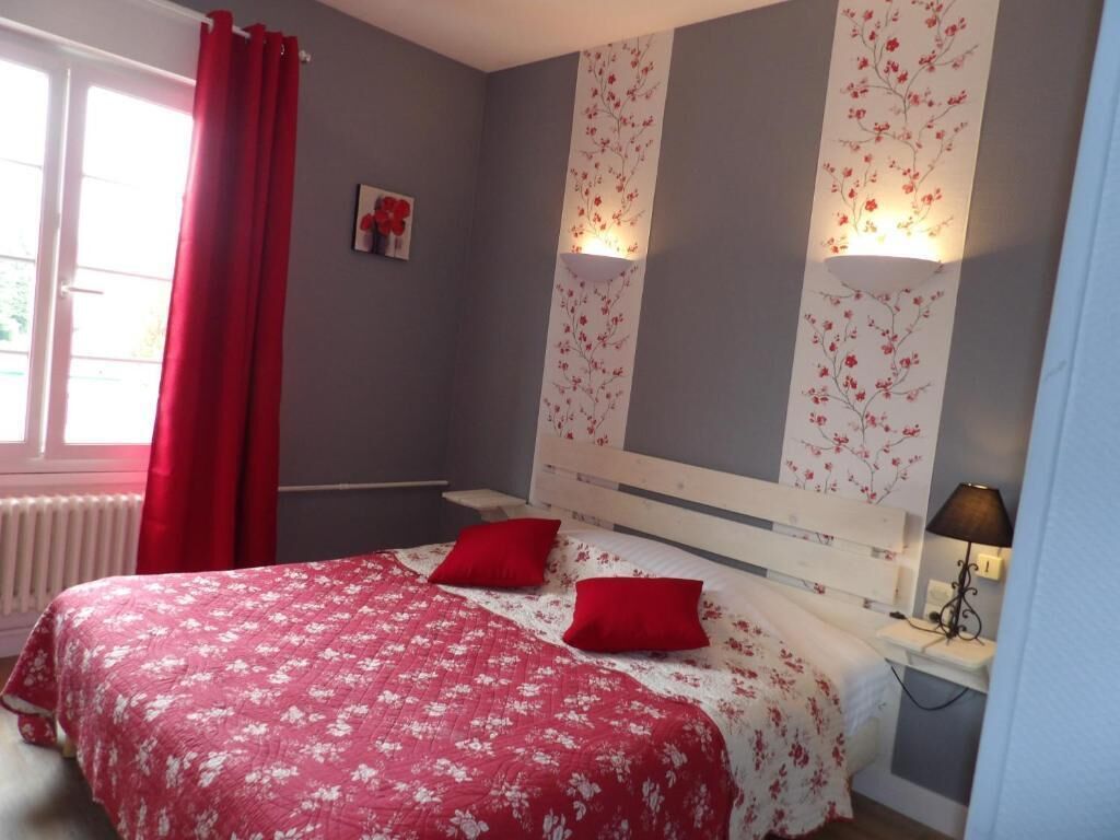 A bedroom with a bed, two red pillows, a nightstand with a lamp, and a window with red curtains.