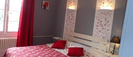 A bedroom with a bed, two red pillows, a nightstand with a lamp, and a window with red curtains.