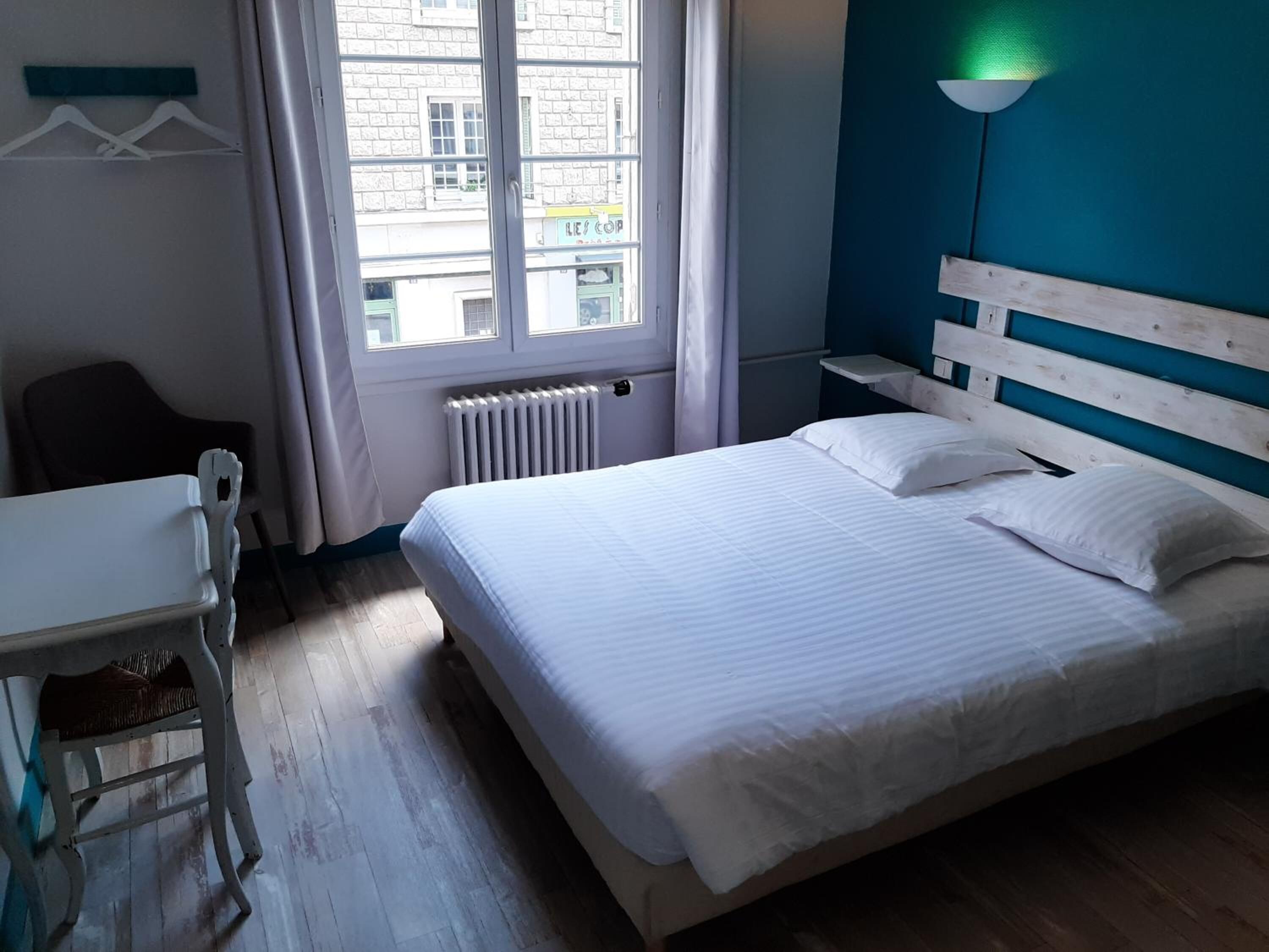 A hotel room with a bed, a desk, a chair, a window, and a radiator.