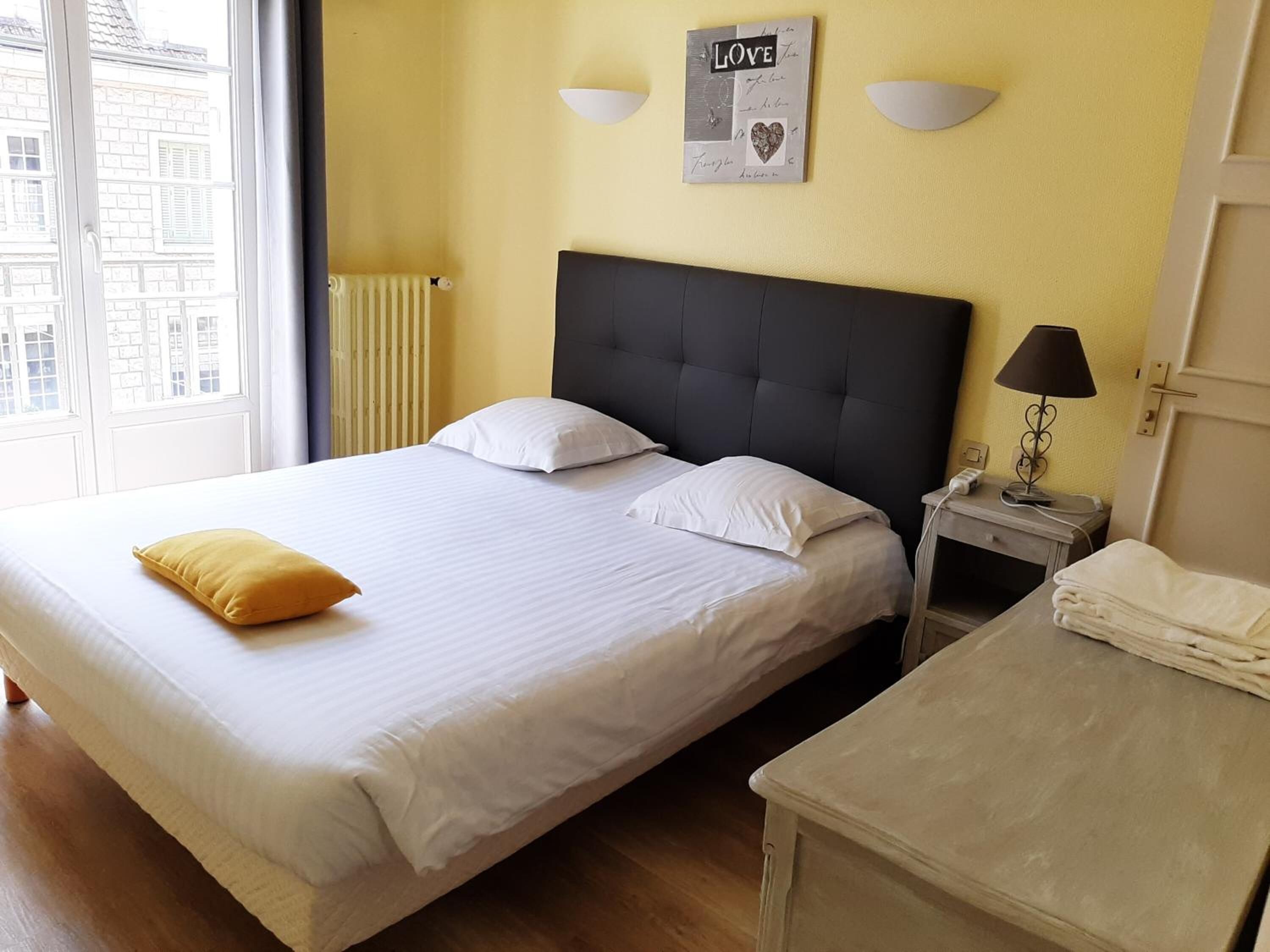 Habitación de hotel con una cama grande, una mesita de noche con lámpara y vistas a edificios a través de la ventana.