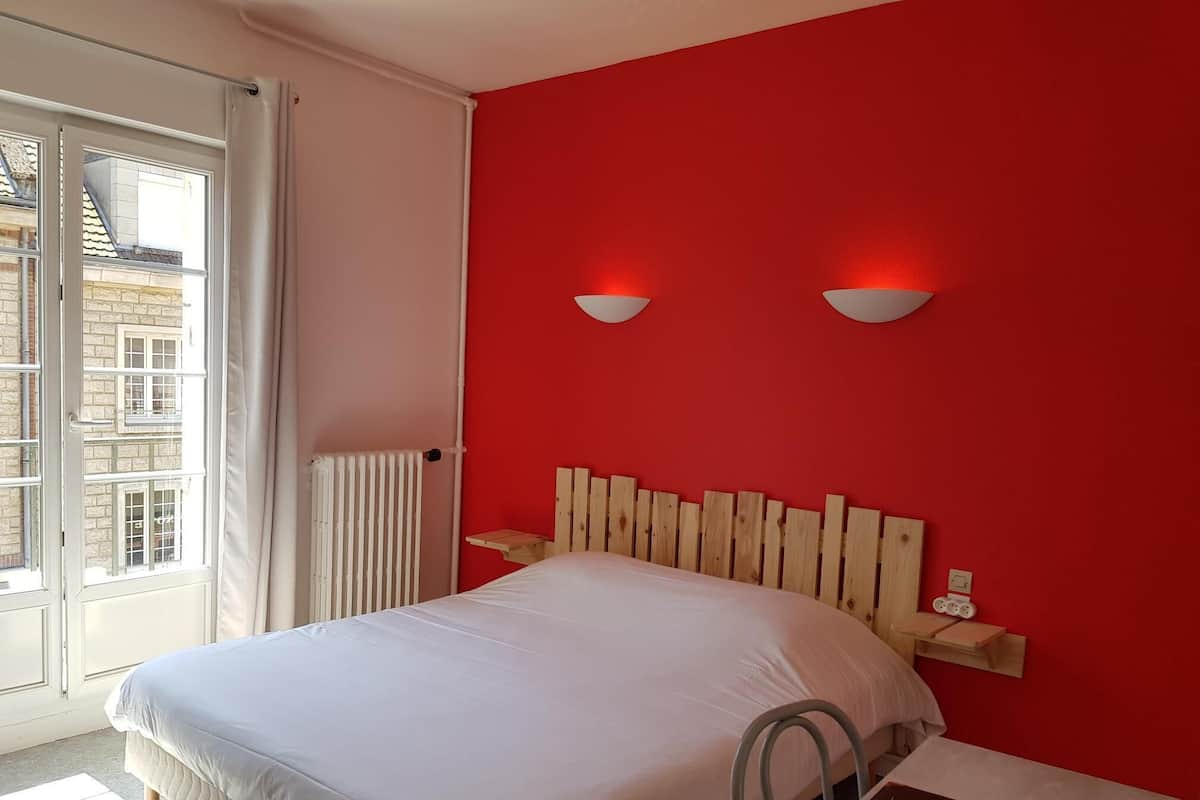 A bedroom with a red wall, a white bed, a wooden headboard, and two wall-mounted light fixtures.