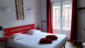 Quarto com cabeceira vermelha, roupa de cama branca, duas almofadas vermelhas, mesa de cabeceira com abajur, luz na parede e janela com cortinas vermelhas.