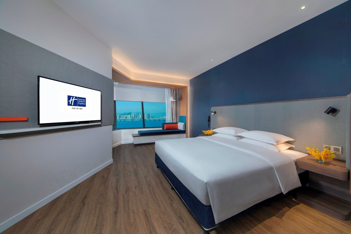 A modern hotel room with a bed, a television on the wall, a window with a city view, and a bedside table with a vase of flowers.