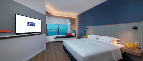 A modern hotel room with a bed, a television on the wall, a window with a city view, and a bedside table with a vase of flowers.