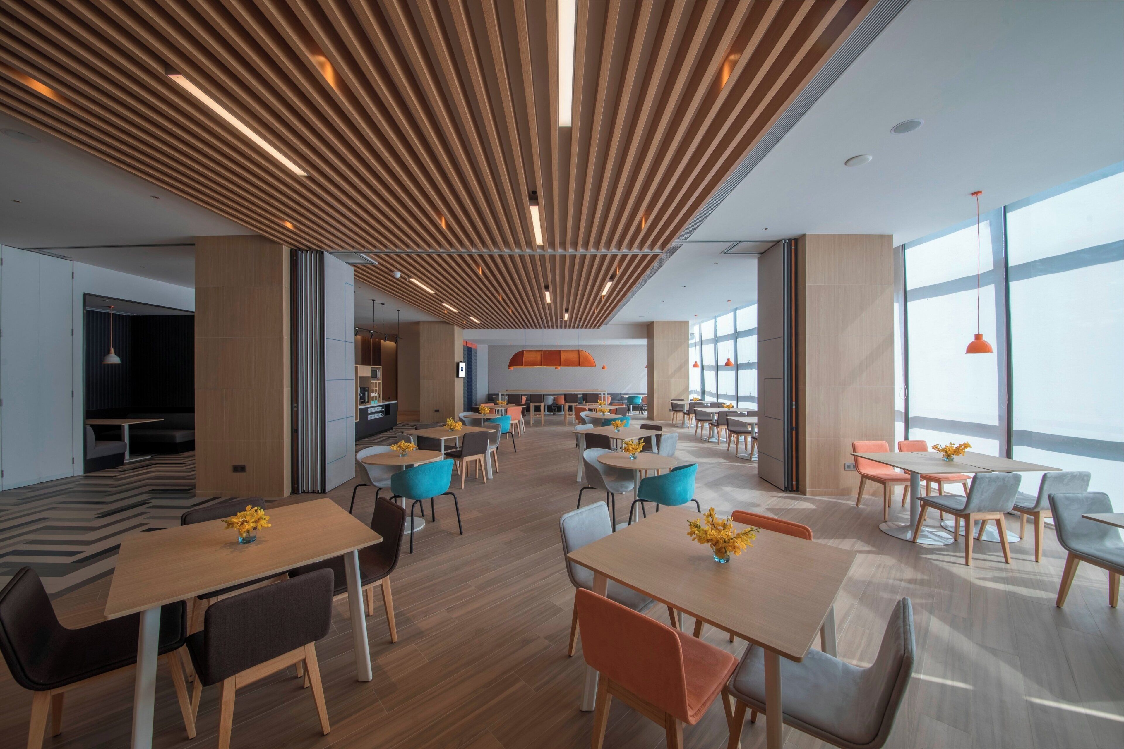 A modern interior with wooden slatted ceilings, multiple seating areas with tables and chairs, and large windows allowing natural light.