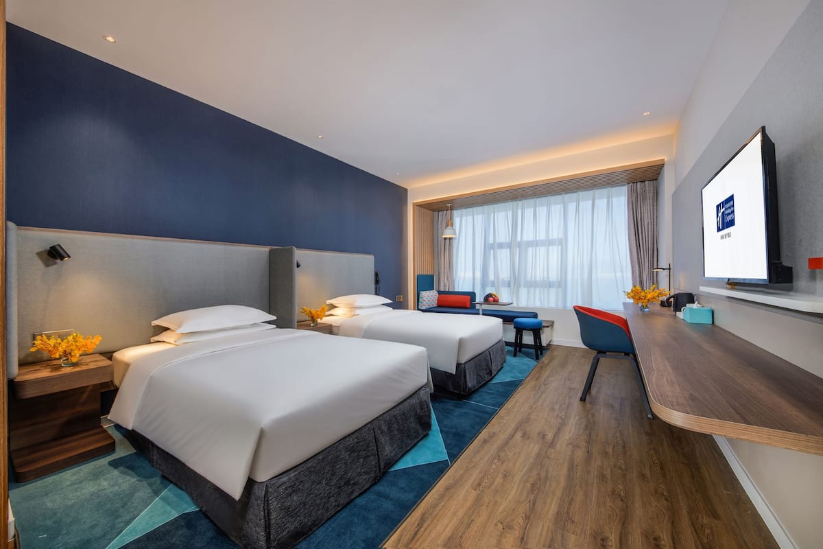 A hotel room with two beds, a desk, a TV, and a window with curtains.