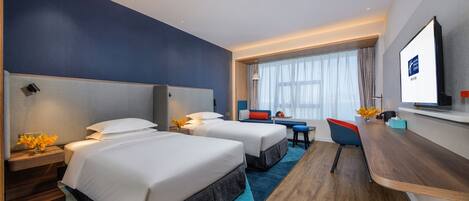 A hotel room with two beds, a desk, a TV, and a window with curtains.