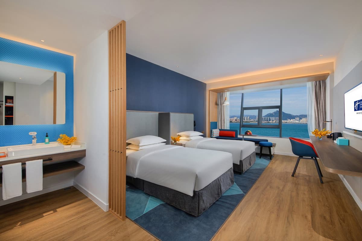 A hotel room with a large window, two beds, a desk, and a bathroom with a mirror and sink.
