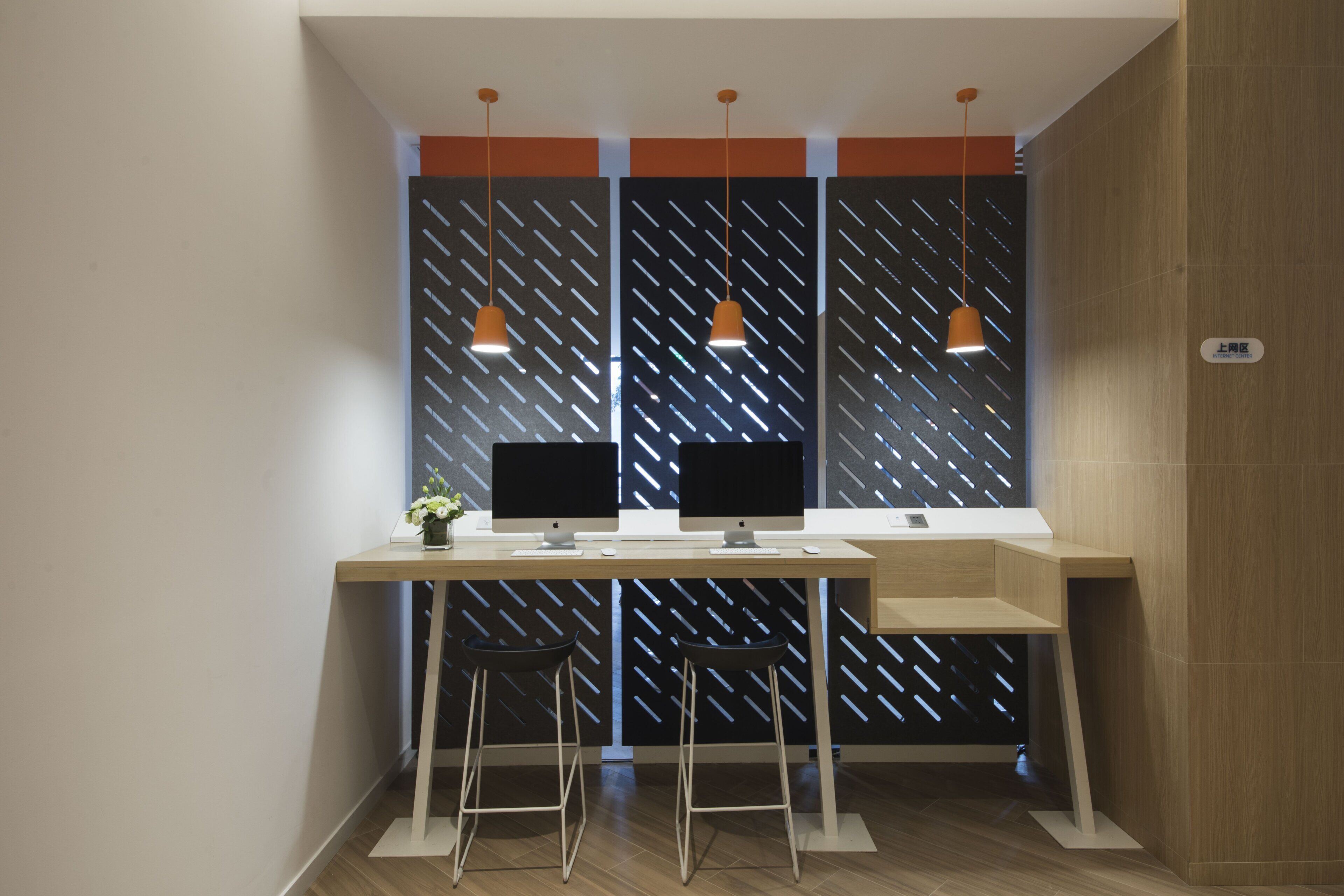 A modern workspace with two desks, each with a computer monitor, keyboard, and mouse. There are two chairs and a small plant on the left desk. The room has wooden flooring and a sloped ceiling with pendant lights.
