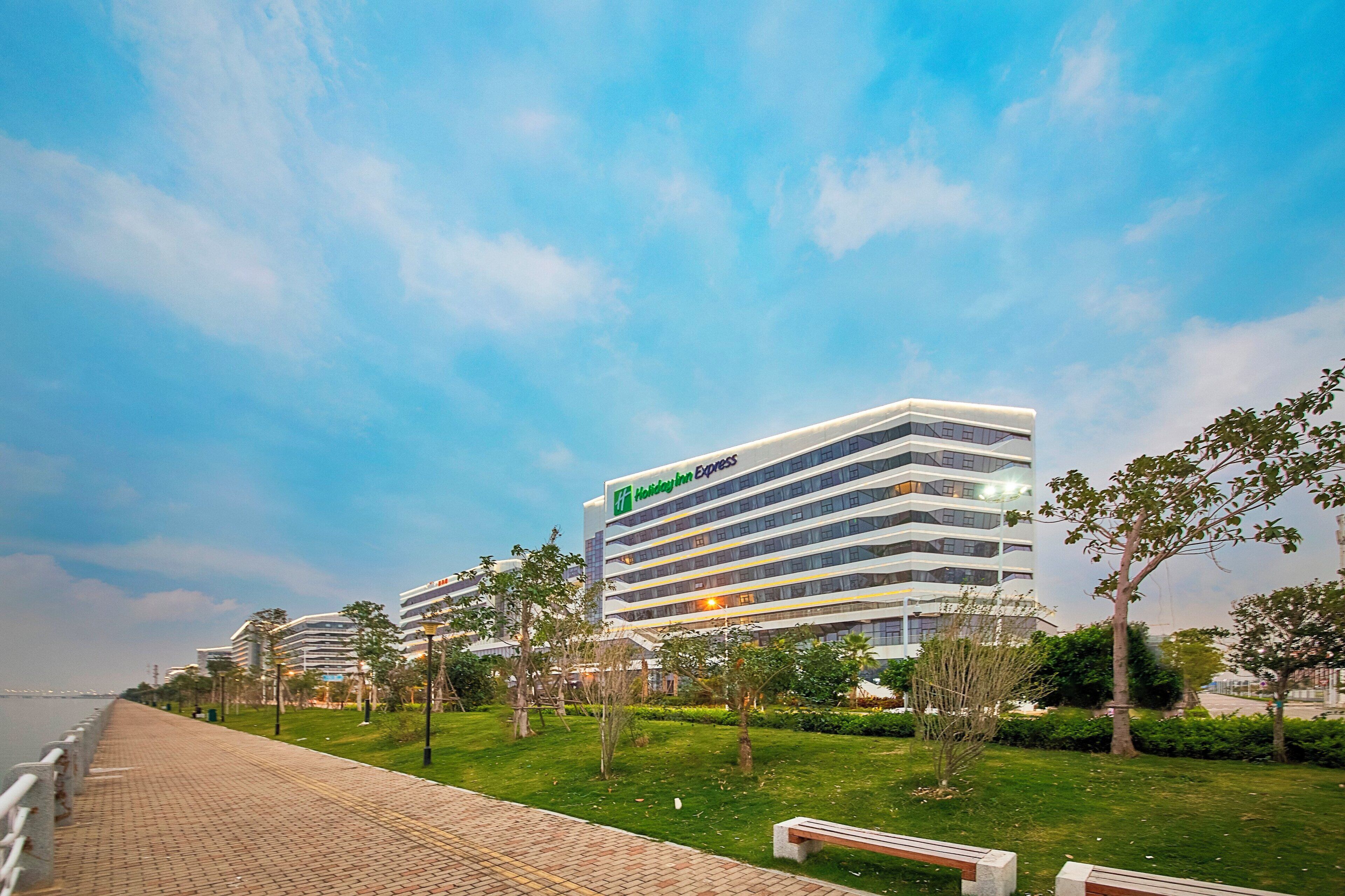 A modern hotel building with a prominent sign, surrounded by a landscaped area with benches and trees.