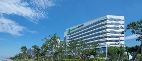 A modern Holiday Inn Express hotel with a glass facade, located near a waterfront with a paved walkway and greenery.