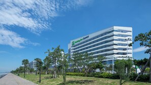 A modern Holiday Inn Express hotel with a glass facade, located near a waterfront with a paved walkway and greenery. - Holiday Inn Express Xiamen Airport Zone by IHG (Xiamen)