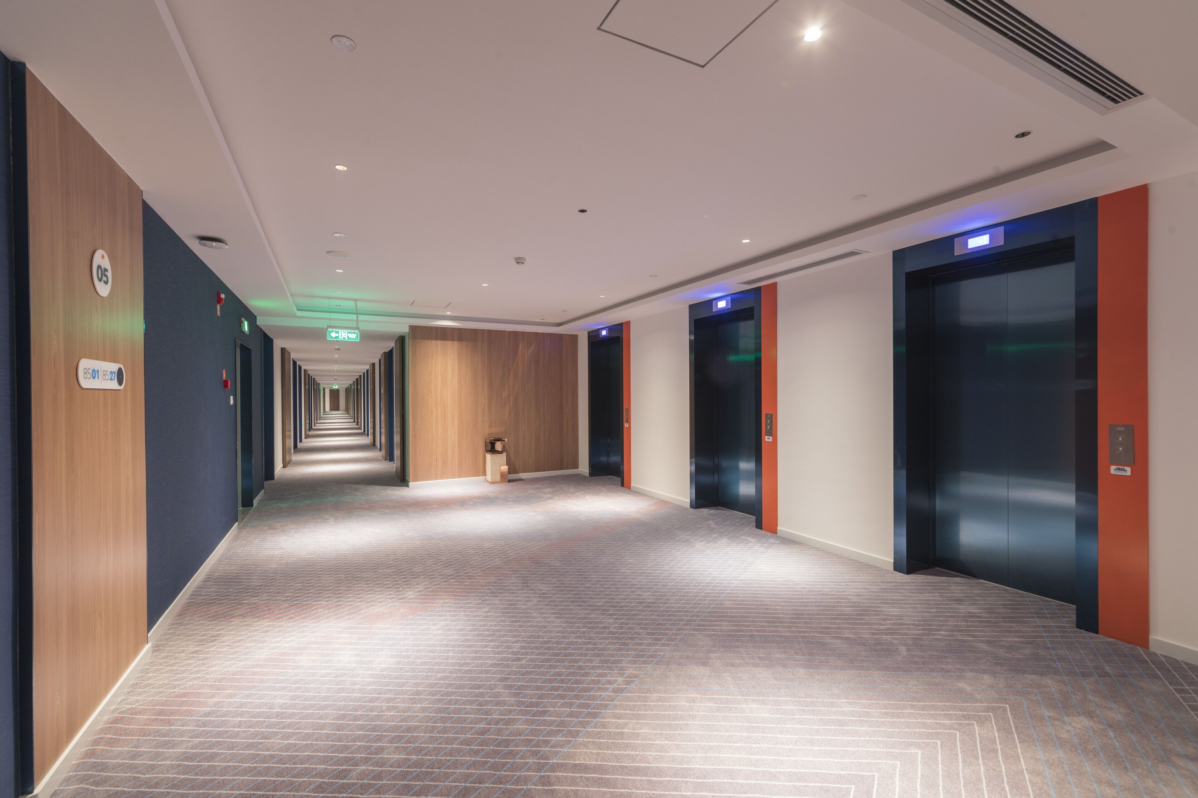 A modern hotel corridor with a herringbone patterned floor, wooden panel walls, and a green emergency exit sign.