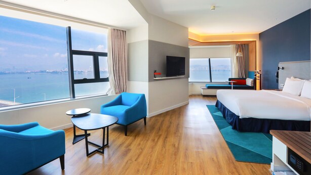 Habitación de hotel moderna con un ventanal grande que ofrece vistas a la ciudad, una cama, un escritorio y una zona de estar con sillas.