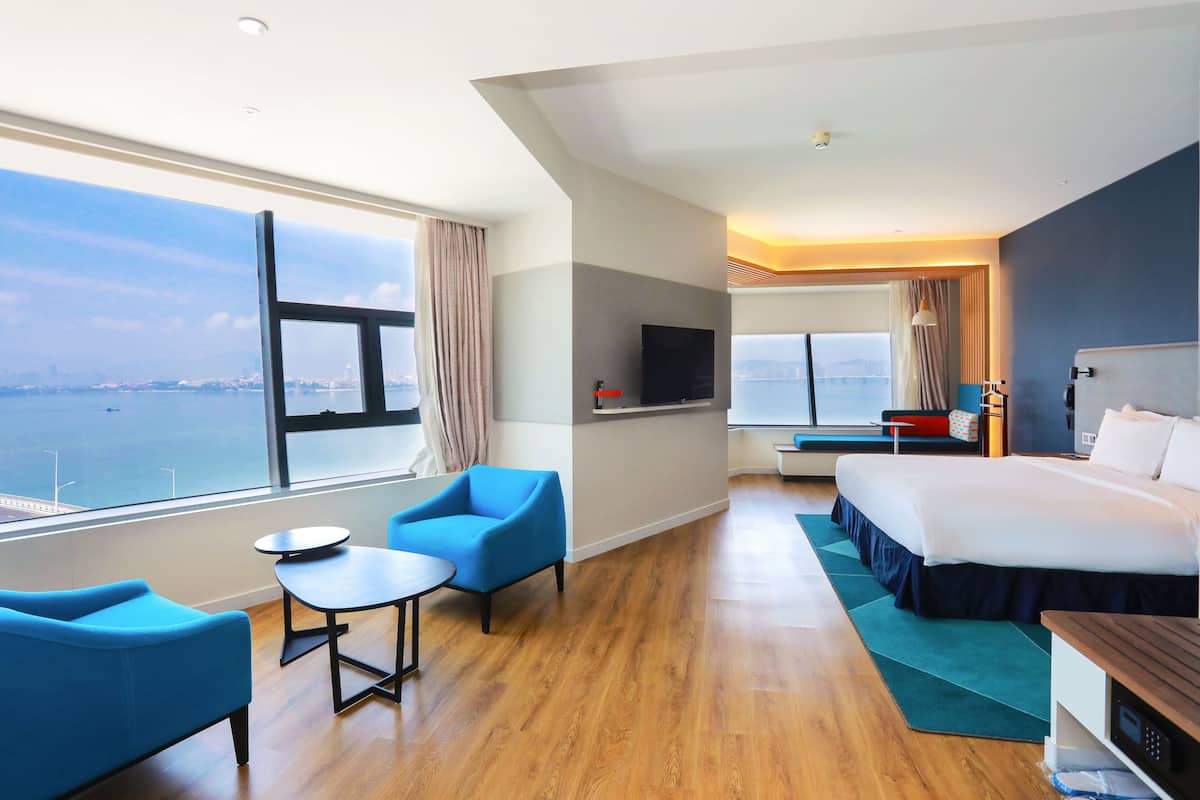 A modern hotel room with a large window offering a city view, a bed, a desk, and a seating area with chairs.