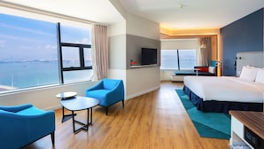 A modern hotel room with a large window offering a city view, a bed, a desk, and a seating area with chairs.