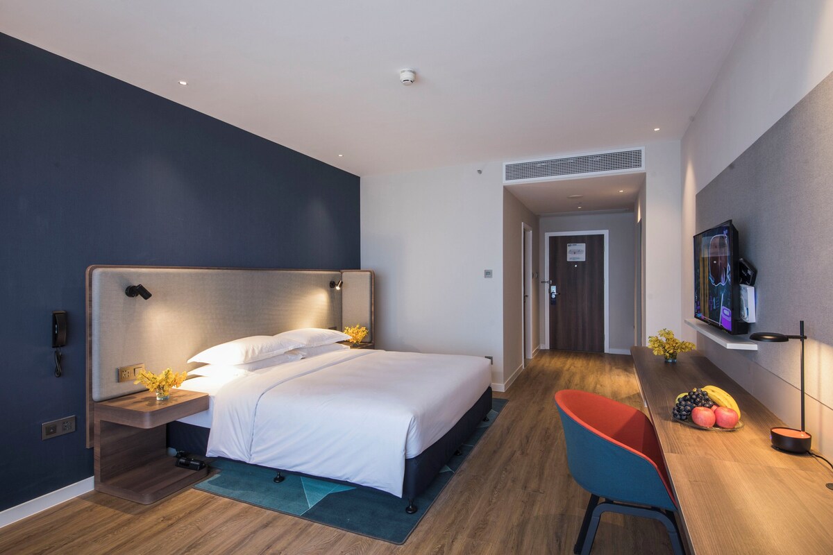 A modern hotel room with a large bed, bedside tables, a desk with a chair, a television, and a fruit bowl.