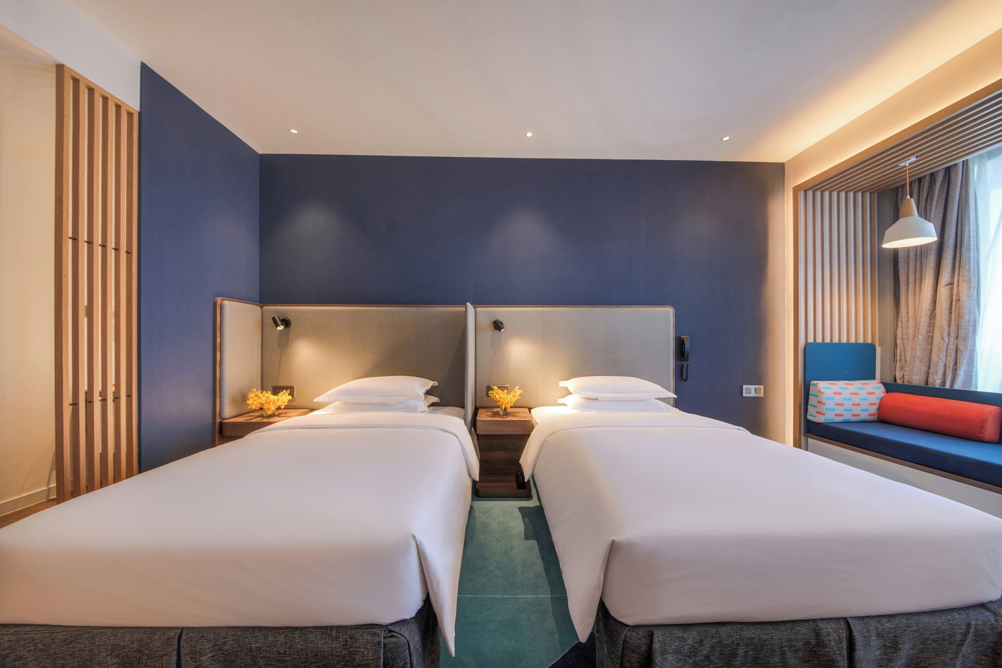 A hotel room with two single beds, a window seat with a red cushion, and a wooden slat wall.