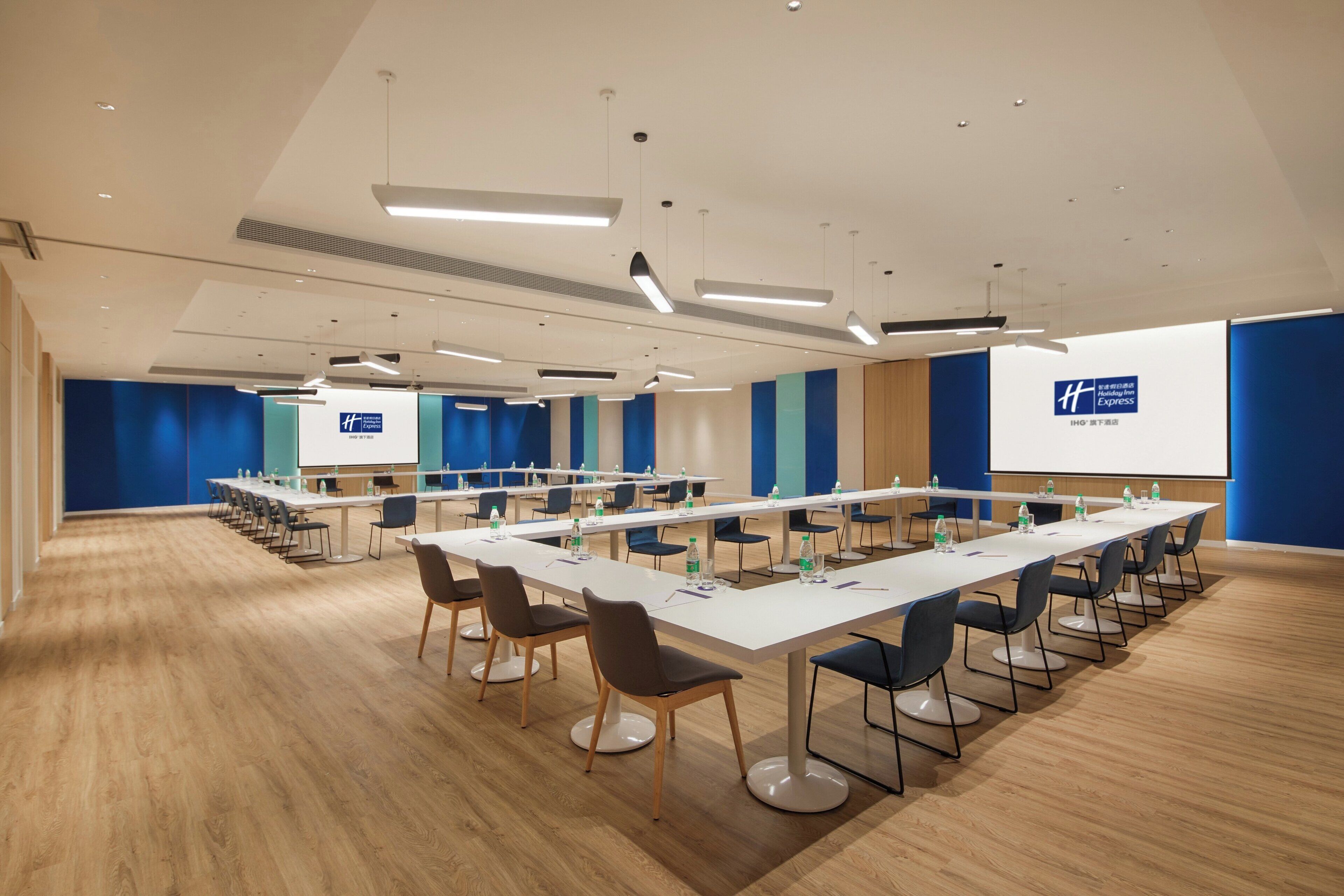 A conference room with long tables, chairs, and a large screen displaying a logo.