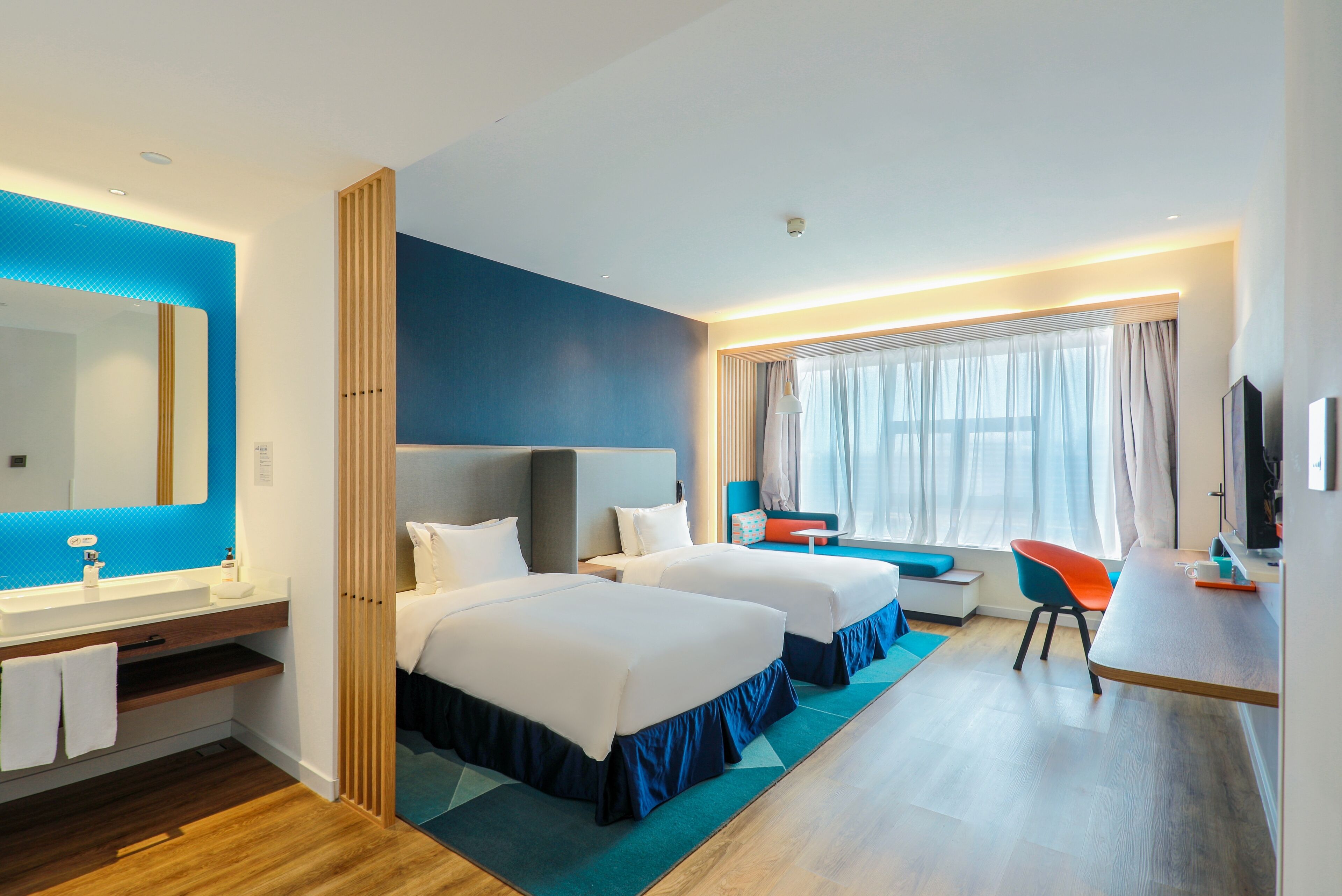 Habitación de hotel con dos camas, un escritorio, una silla, un televisor y un baño con espejo y lavamanos.