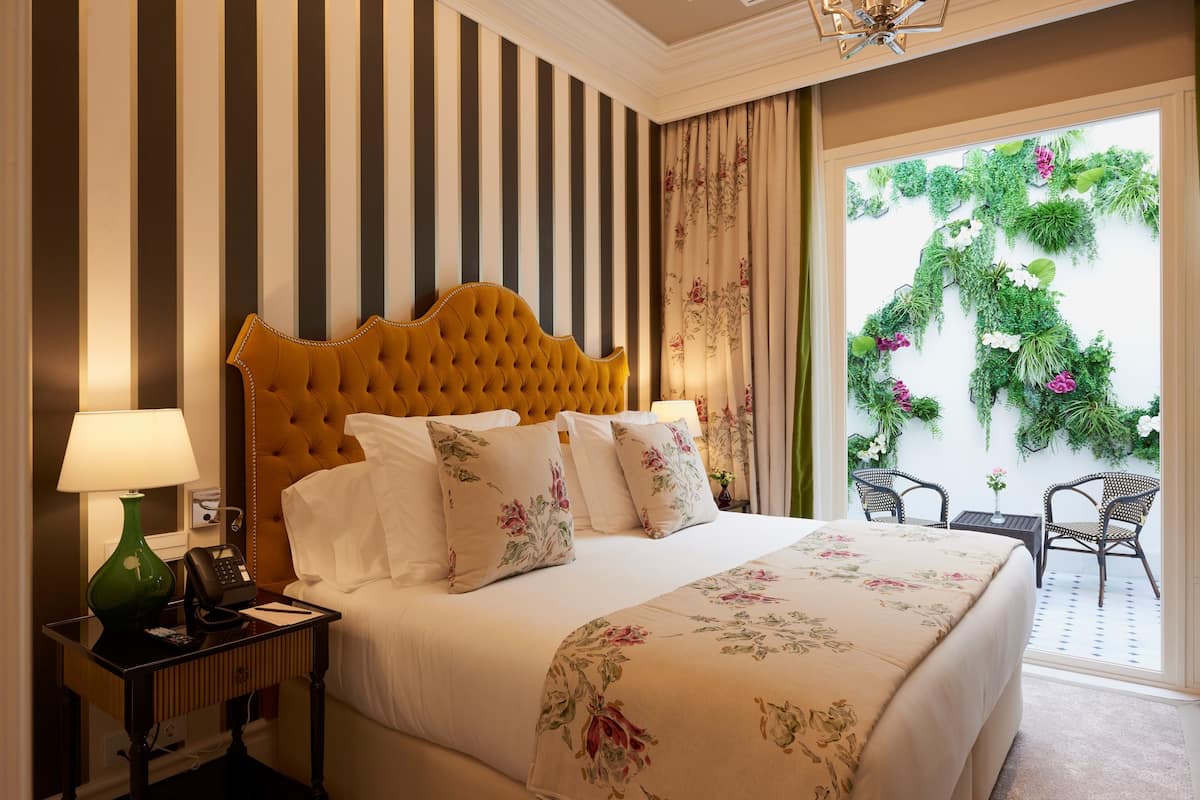 A hotel room with a large bed, striped walls, a green lamp, a telephone, and a view of greenery through the window.