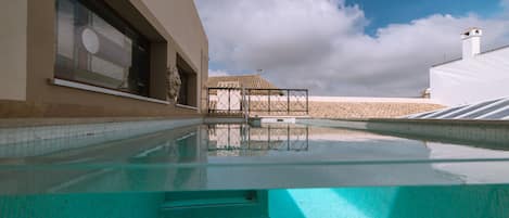 Una piscina con agua cristalina, un suelo de baldosas y un edificio con ventanales grandes y techo de tejas al fondo.