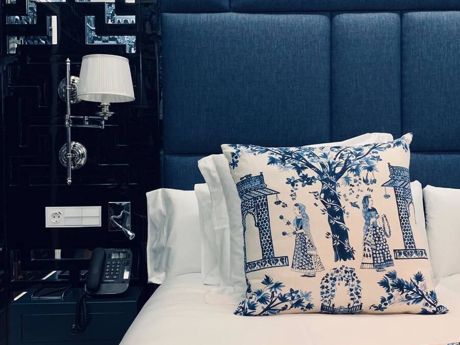 Ett hotellrum med en blå, klädd sänggavel, en säng med vita och blå blommig sängkläder, ett svart nattduksbord med en lampa och en telefon.