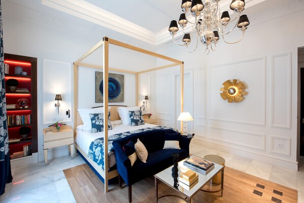 Una cama con dosel, cabecera tapizada en azul, una mesita de noche con lámpara, una estantería roja y un espejo redondo dorado en la pared.