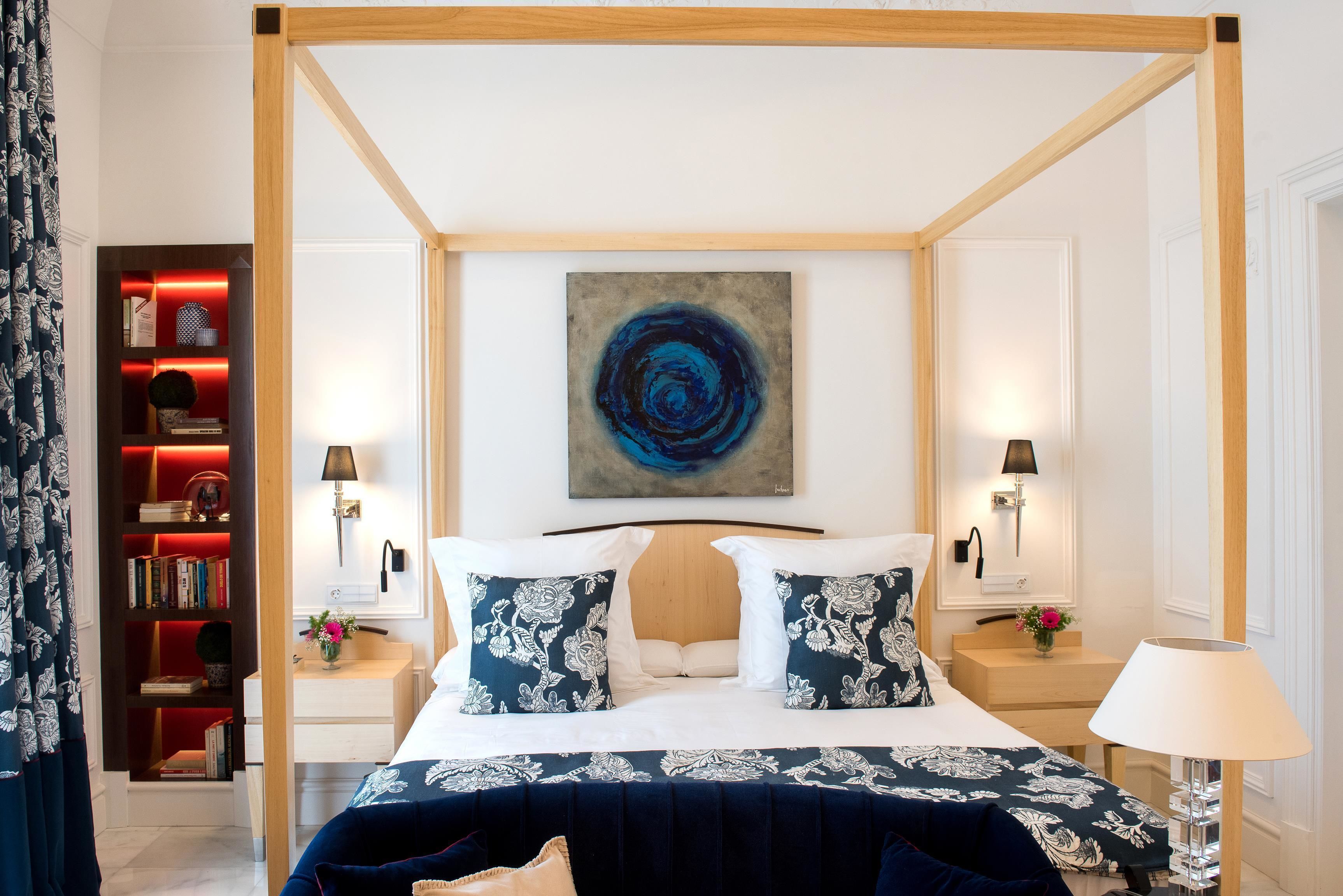 A four-poster bed with a dark blue and white floral bedspread, two matching pillows, and a modern abstract painting above.