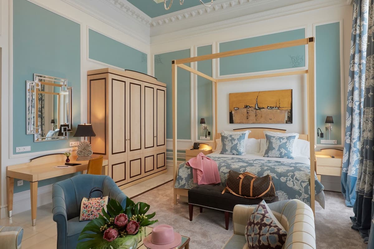 A spacious room with a large bed, a sitting area with a sofa and armchairs, a desk with a chair, a wardrobe, and a chandelier.