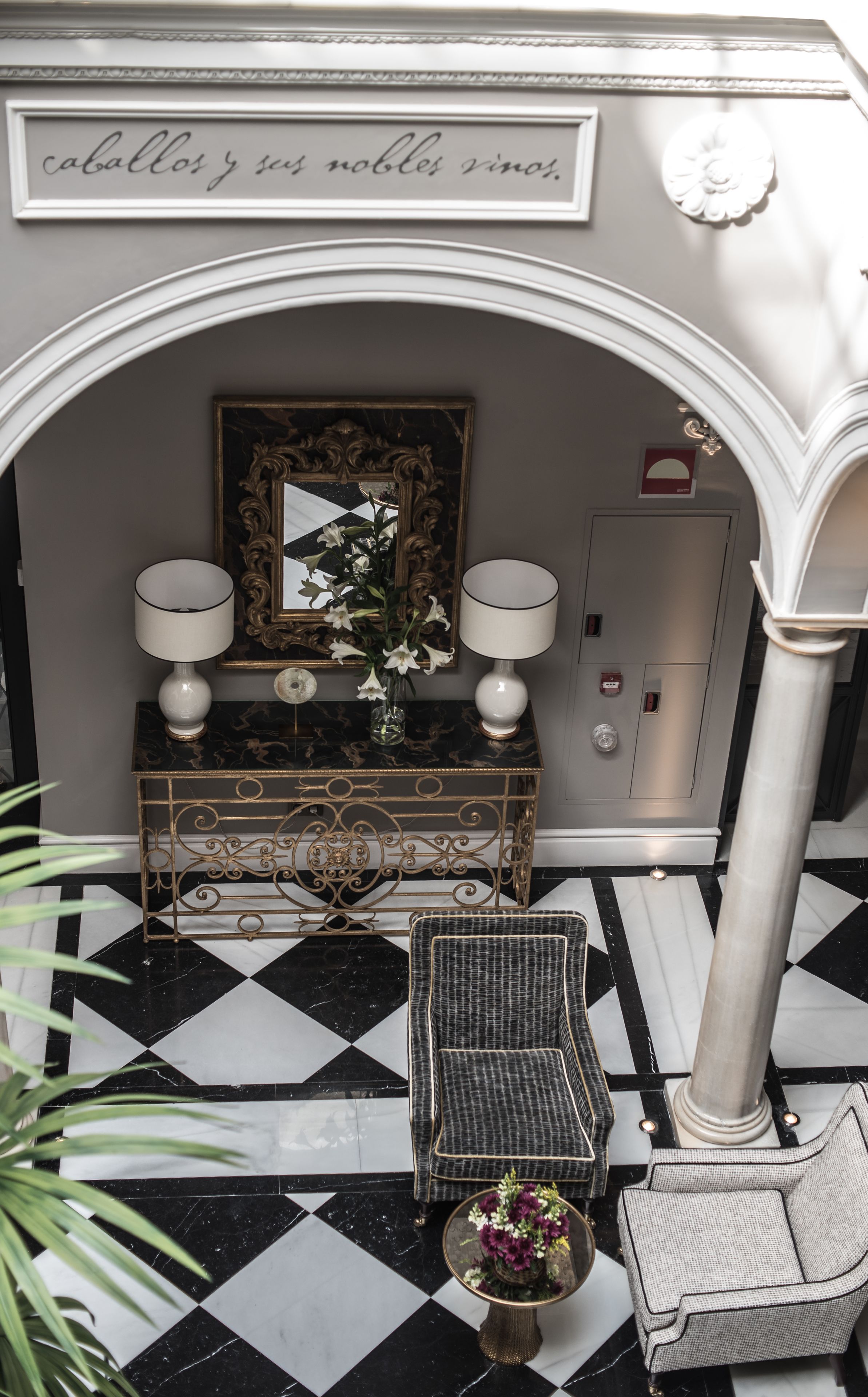 A black and white checkered floor, a mirror, two lamps, a vase with flowers, a wicker chair, and a sign above the mirror.