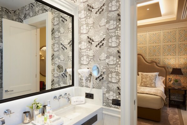 Un baño con un espejo grande, un lavabo y un mueble de tocador. Se observa una cama con cabecera y una mesita de noche con lámpara.