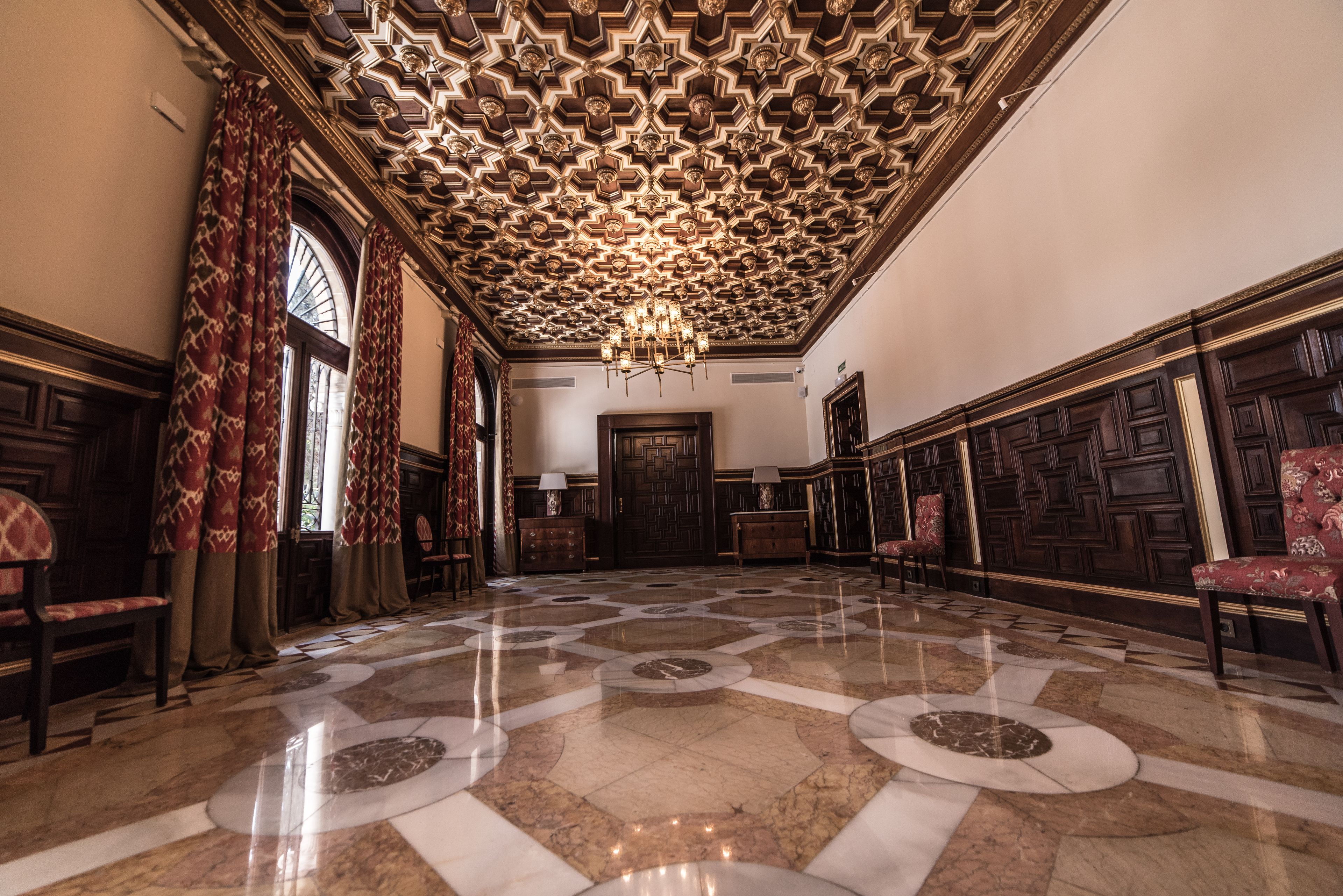 Un salón imponente con techo ornamentado, grandes ventanales con cortinas rojas y un suelo de mármol pulido.
