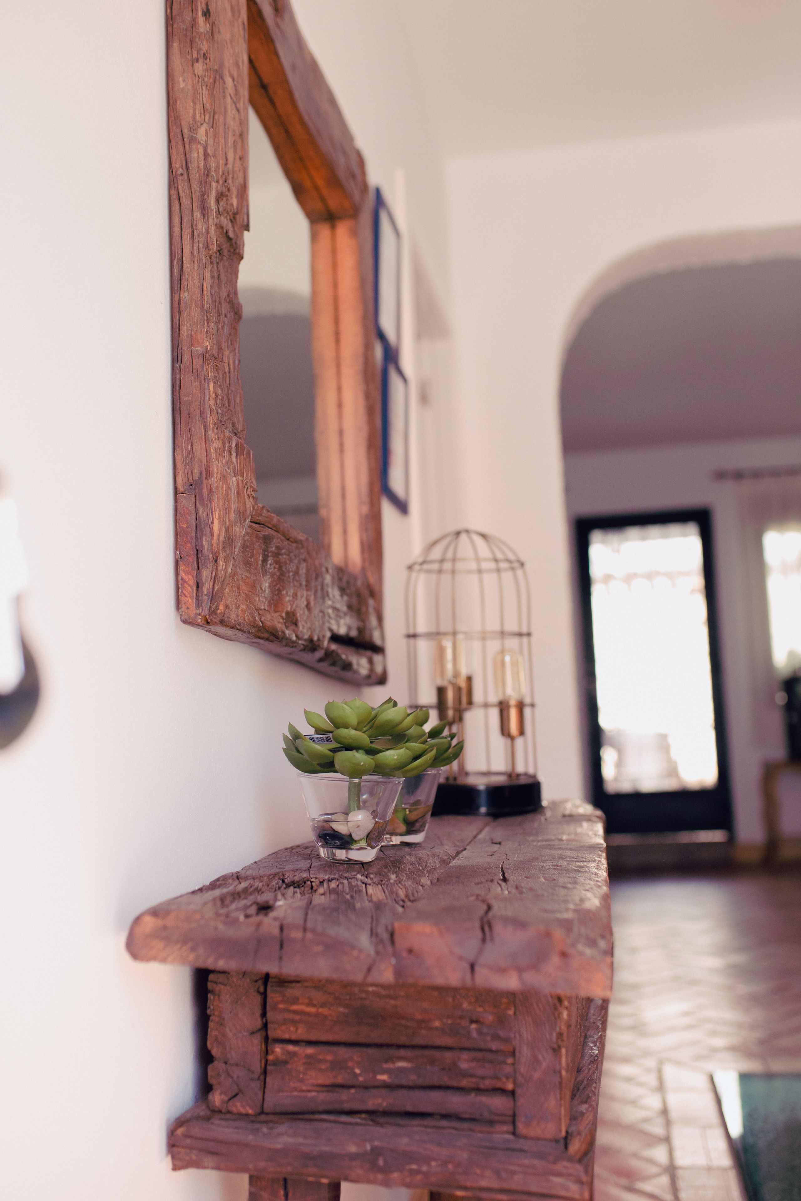 Una mesa consola de madera rústica con una suculenta en maceta y un florero de vidrio. Una jaula de pájaros de estilo vintage con luces está sobre la mesa.