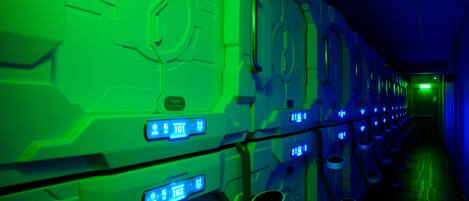 Un pasillo con filas de cápsulas futuristas iluminadas en verde, cada una con una pantalla digital que muestra números.
