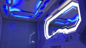 Ein futuristisches Zimmer mit einem Bett, einer Bedienkonsole und einem Fenster mit blauer Beleuchtung.