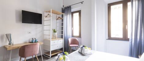 A modern hotel room with a wooden desk, a pink chair, a bed with towels and flowers, a TV, and a window with curtains.