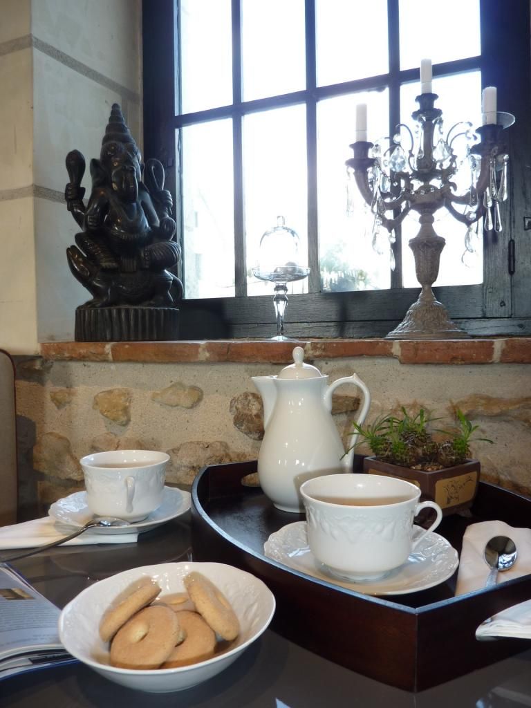 A table with a tea set, a bowl of cookies, and a window with a decorative statue.