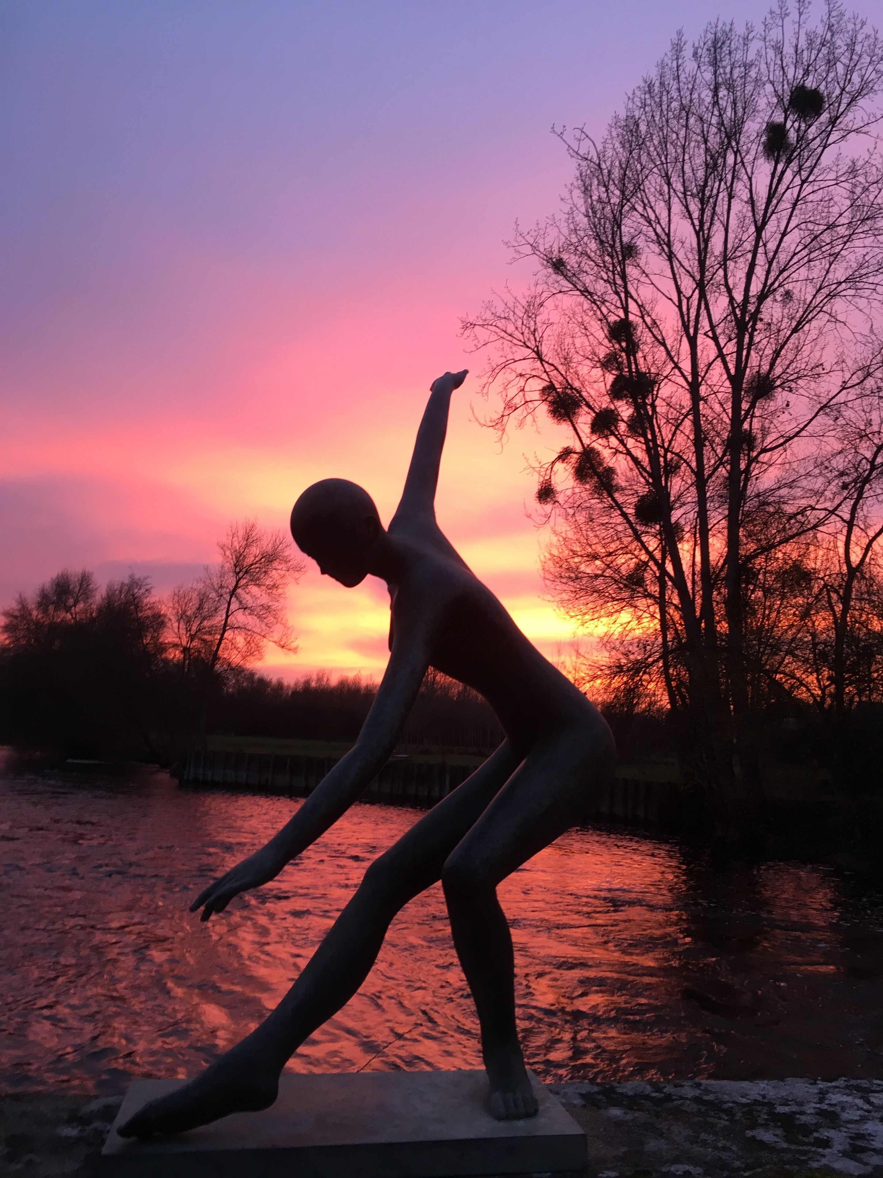Una statua raffigura una persona in posa dinamica, con un corpo d'acqua e un tramonto sullo sfondo.
