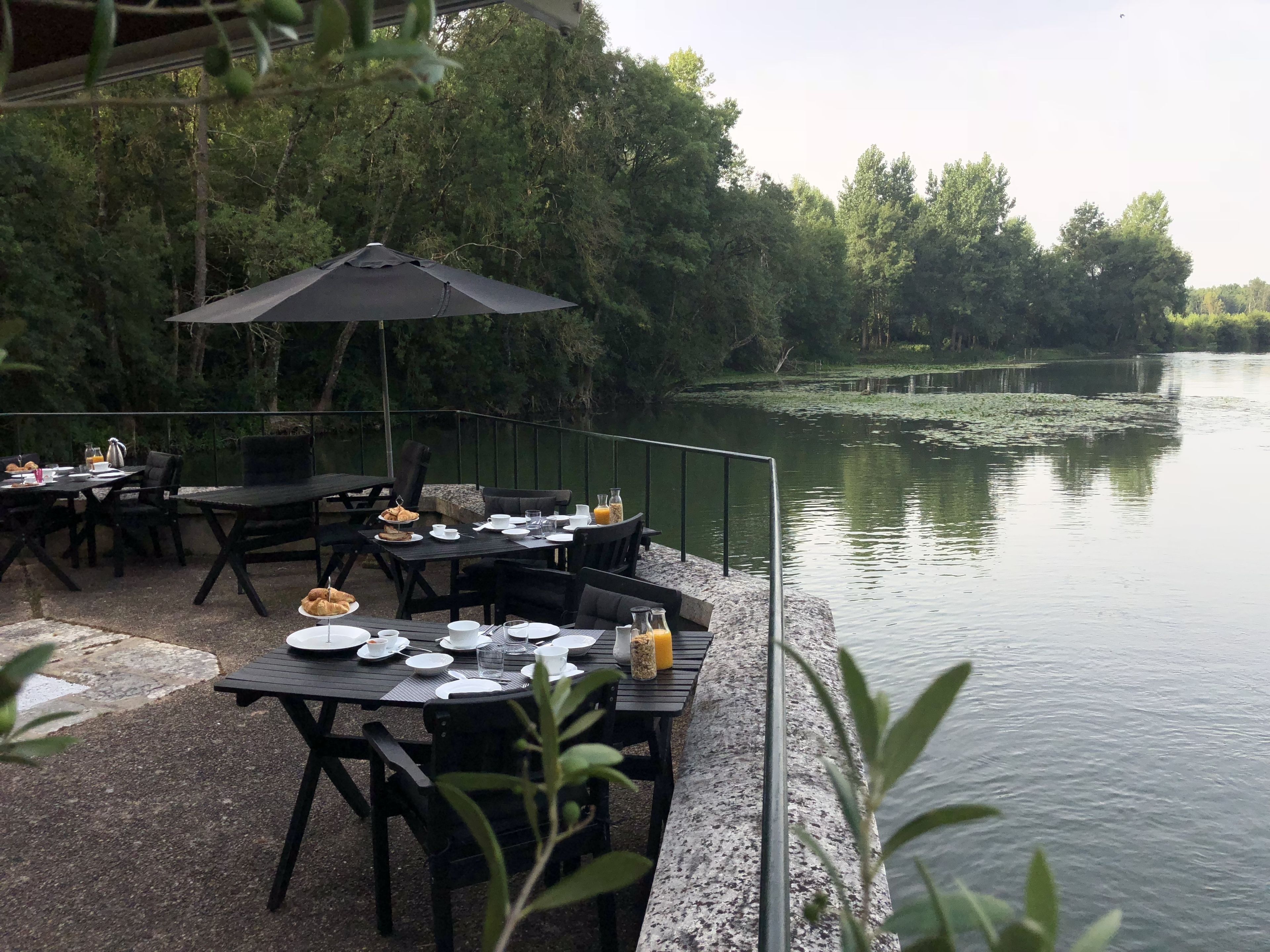 An outdoor dining area with tables and chairs, a black umbrella, and a view of a calm lake surrounded by trees.