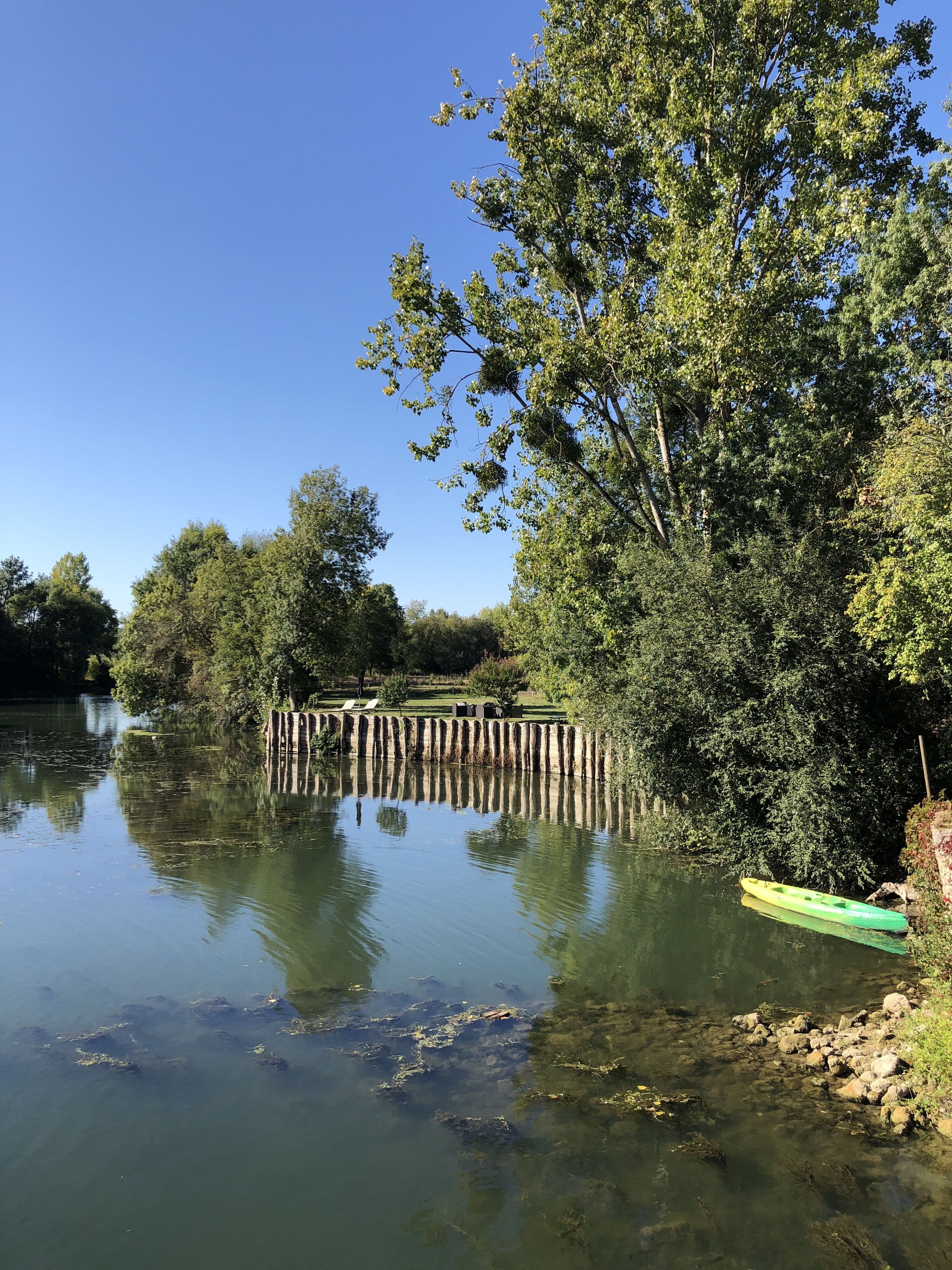 A calm river with a green kayak on the right bank, surrounded by dense trees and a clear blue sky.
