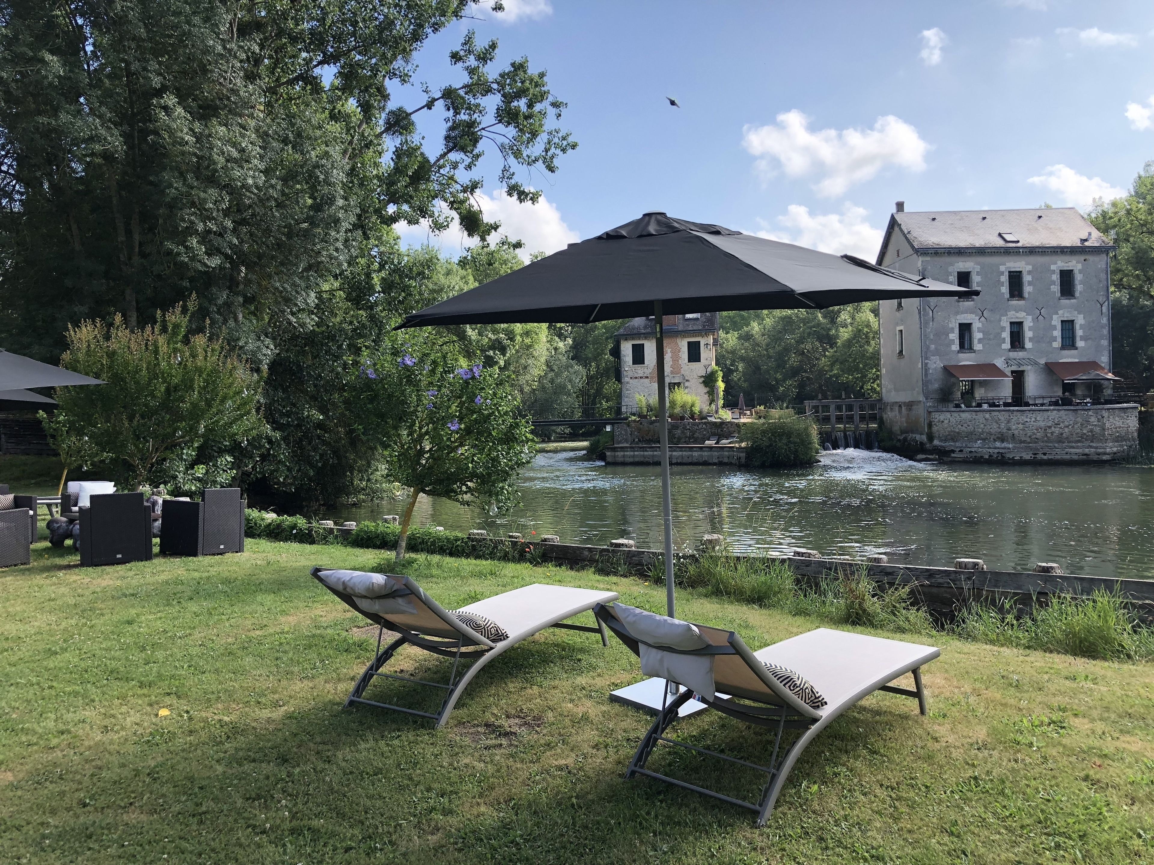 Un patio junto al río con dos sillones de relax y una gran sombrilla, con vistas a un edificio histórico y un río tranquilo.